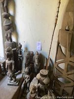 Overview of multiple carved wood sculptures lined against wall, including totem, figurines, panel, staff, drum