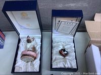 Two navy satin-lined presentation boxes each holding a feline enamel trinket box: Siamese Kitten on pink base and a curled cat in basket.