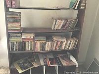 Wooden bookshelf with three shelves holding stacks of paperback books on upper two shelves and vinyl records on lower shelf