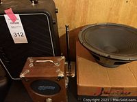 Three speakers in used condition including a Pignose 7-100 amplifier, a large round speaker on a box, and a Vox speaker cabinet