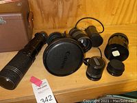 Wide view of three lenses on wooden shelf: long telephoto lens, servo zoom lens with hood and grey zoom lens, plus extension tubes and adapter ring