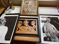 Four framed artworks displayed on tabletop: Western painting, wood relief, two Zappa photographs
