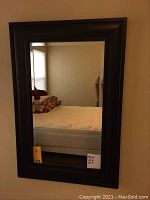Front view of rectangular beveled wall mirror with dark wood frame hanging on a wall, showing reflection of bed and pillows.