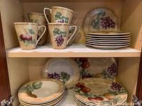 Shelf view of dish set showing stacked plates, bowls and 7 mugs with fruit motifs including grapes and cherries.