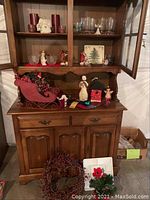 Full view of wooden hutch and cabinet loaded with assorted Christmas decorations including red sleigh, figurines, glass candle holders, birdhouse, and wreath.