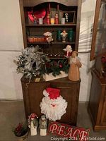 Assorted Christmas decorations displayed on and around a wooden cabinet including a glittery wreath, multiple figurines, greenery, candles, and a PEACE pillow.