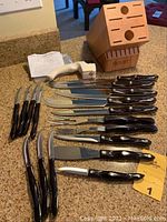 Overhead view of wooden block, white pull-through sharpener, large knives, forks, spatula, paring knife, and cluster of steak knives arranged on countertop.