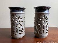 Pair of cylindrical ceramic candlestick holders with beige and dark brown glaze, featuring cut-out floral design shown from side.