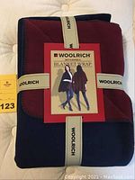 Folded Woolrich reversible blanket wrap with navy and cabernet colors, secured with branded ribbons, showing packaging label with model image.