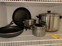Wide view of all pots and pans included in the lot on a wire shelf, showing the large Revere Ware pot with lid, two non-stick fry pans, and two small saucepans.