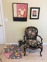Photo showing Queen Anne style chair with large floral upholstery, reversible floral apron, white pitcher with artificial pink peonies, and dishes set near the chair with floral artworks on the wall behind.