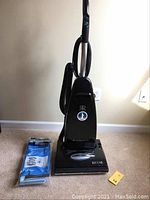 Full frontal view of Riccar Vibrance upright vacuum with attached hose and handle, placed on carpeted floor with vacuum bags beside it.