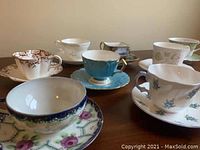 Seven different tea cups with matching saucers displayed on a wooden table. Various floral and colored patterns visible.