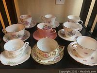 Eight vintage porcelain tea cup and saucer sets with floral designs and gold accents arranged on a shelf.