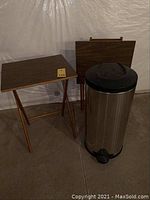 Two wood tray tables with medium brown woodgrain finish next to a cylindrical metal trash bin with black lid and foot pedal, interior corner setting.