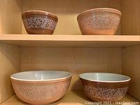 All four Pyrex Woodland mixing bowls shown on wooden shelf, two on top shelf and two on bottom shelf, displaying brown floral pattern exterior.