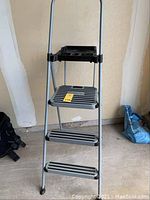 Frontal view of the ladder showing three wide steps with black anti-slip strips, the work tray on top, and the steel frame