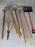 Photo showing the full set of 7 long handled yard and garden tools laid on concrete, including wooden handled rakes, shovel, garden rake, snow shovel, pruning shears, and edger.