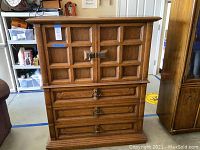 Full front view of the wooden armoire dresser showing the paneled cabinet doors and three drawers with metal pulls. The cabinet doors have decorative square panels and are latched with a metal fixture.