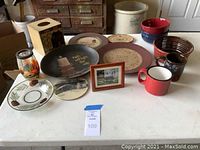 Photo of entire lot showing plates, mugs, bowls, tissue holder, lantern and framed art on a white table