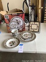 View of Gibson Christmas Toile 16-piece dinnerware set in original box with some items displayed on a table. Also shown is a metal tree candle holder and framed sign that reads 'Friends gather here'.