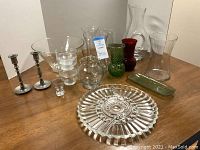 Overview of glass serving tray, glass salad bowl, assorted clear, green and red glass vases, and silverplate candlesticks on a wooden table.