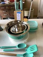 Photo showing knife block with turquoise handle knives, mixing bowls stacked with turquoise bases, and plastic storage containers with turquoise lids along with turquoise cooking utensils on a table.