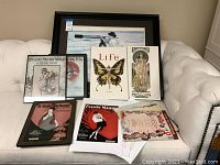 Full view of framed fishing print and assortment of sheet music cover prints, some framed, some loose on a white couch.