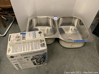 Photo showing stainless steel double bowl kitchen sink and boxed InSinkErator garbage disposal side by side on floor.