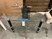 Photo showing the full TV stand with three clear glass shelves and black legs with silver metal accents.