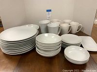 Full overview of the assortment of white dishes including stacks of dinner plates, small plates, bowls, and a collection of mugs arranged behind them.