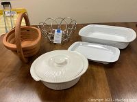 Photo showing five items on a wooden table: two white ceramic Corningware casseroles, one with lid, one rectangular; a collapsed wood basket; and a round metal wire basket.