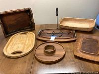 Photo showing assortment of seven items: three wooden boards, two woven basket trays, a wooden tray with metal handles, and a sharpening tool with black handle on a wood table.