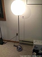 Full view of the floor lamp lit in a room, showing white globe light shade atop chrome pole and hemisphere base.