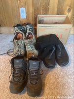 Overall view showing three pairs of hiking boots and a box holding belts with buckles arranged on carpeted floor against wood paneling