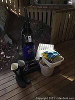 Hoover upright vacuum, black Tretorn rubber boots, white plastic stepping stool, wash bucket with gloves on top placed near a wooden lattice fence.