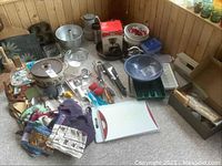 Overview of various pans, bowls, kitchen utensils, plastic trays, cutting boards, aprons, and other kitchen accessories on carpeted floor against wooden wall