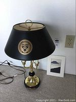 Table lamp with black and brass design, featuring a gold University of Pennsylvania emblem on lampshade, standing on floor next to framed art.