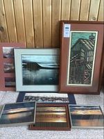 Set of six framed artworks and photographs displayed leaning against a wood wall. Includes a tall vertical framed art print of a coal bunker, several framed photographs with maritime sunset and boat themes lying flat or leaning.