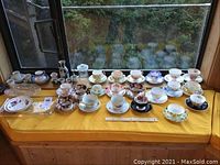 Wide view showing a large selection of teacups with matching saucers arranged on yellow cloth with sake set and glass plates visible