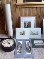 View showing a black metal double-sided clock and six framed or matted travel art prints arranged on carpet with a wooden wall background.