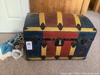 Photos of a vintage dome-top trunk with a painted exterior featuring bright yellow vertical stripes separating red and blue panels, heavily worn with stains and marks, showing iron hardware and two fabric wind socks beside it.