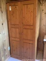 Front view of standalone wood wardrobe showing two paneled doors with round wooden knobs against wood-paneled walls.