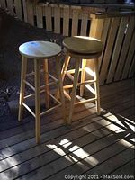 Two natural wood bar stools side by side on a wooden floor, one with swivel seat larger diameter, the other smaller diameter fixed seat stool.