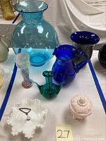 Wide view of all art glass items including blue pitcher, blue and teal vases, milk glass candy dish and jar on white cloth background.