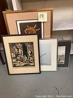 Four framed art pieces leaning against a wall, depicting various scenes including an abstract sun, black and white woodcut print, and architectural photo.