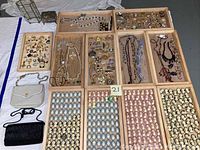 Wide shot showing several wooden trays laid out with assorted costume jewelry including brooches, earrings, necklaces, and cameo pieces. Also shows two small fabric handbags in white and black beside the trays, and a silverplate vanity box in background.