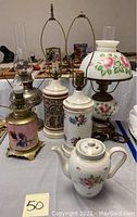Lot display on table showing multiple vintage lamps with floral porcelain bases and the Thomas Bavaria tea pot in front.