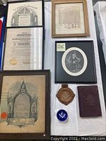 Overview photo showing multiple framed membership certificates, framed print, brown ashtray, blue glass paperweight, and a small book on a white cloth.