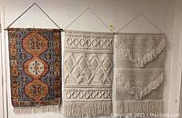 Three wall tapestries hanging side by side on a wall; one is colorful woven and two are cream macramé style